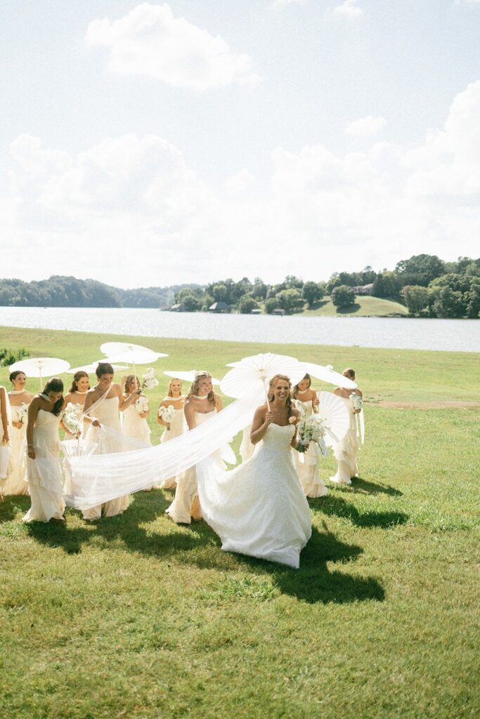 bride and bridesmaids walking across a lakeside lawn with white parasols and flowing veil at an outdoor Nashville TN wedding
