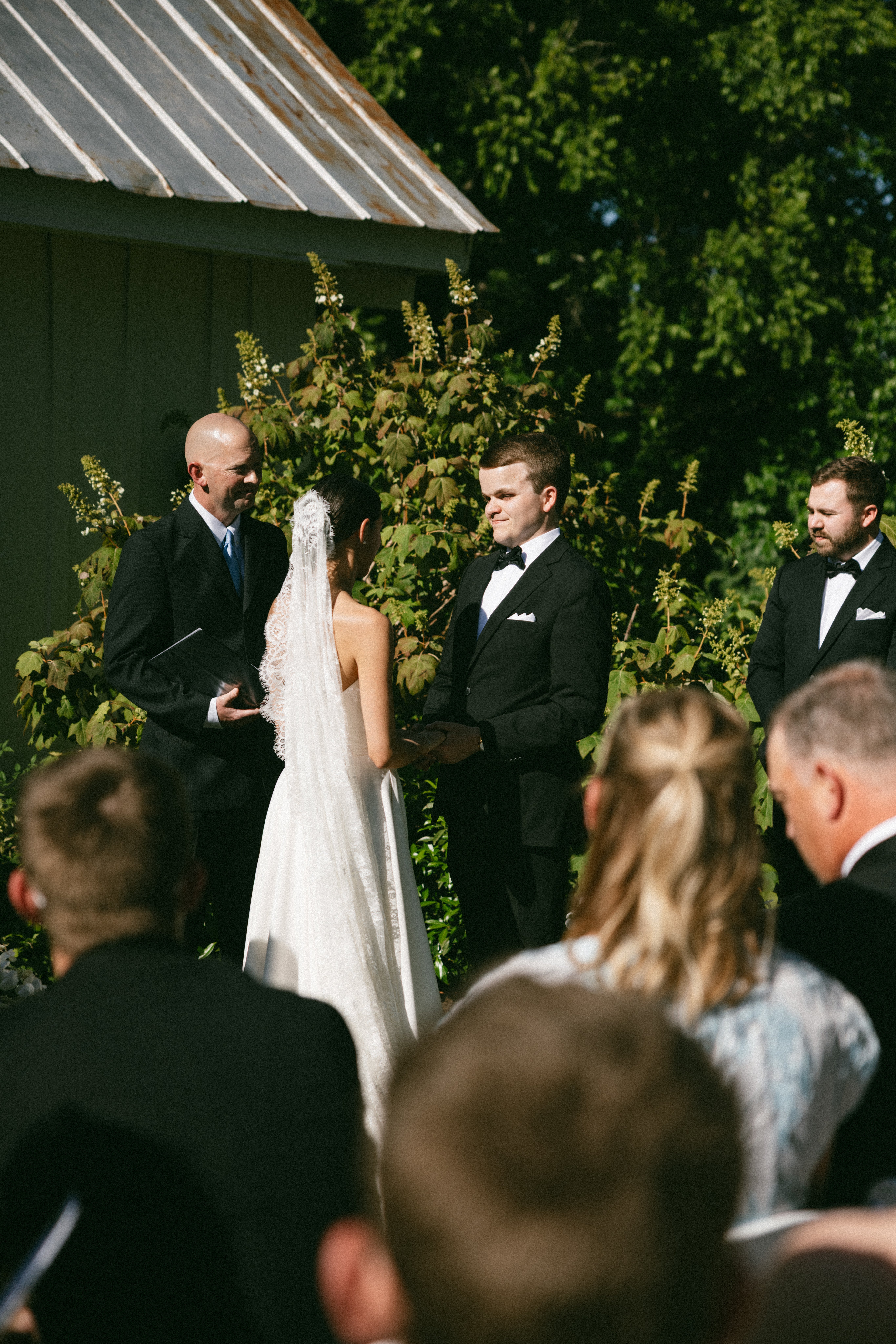 Bride and groom holding hands during an outdoor ceremony in front of a rustic white chapel and garden greenery at one of the most charming small wedding venues near Nashville TN.