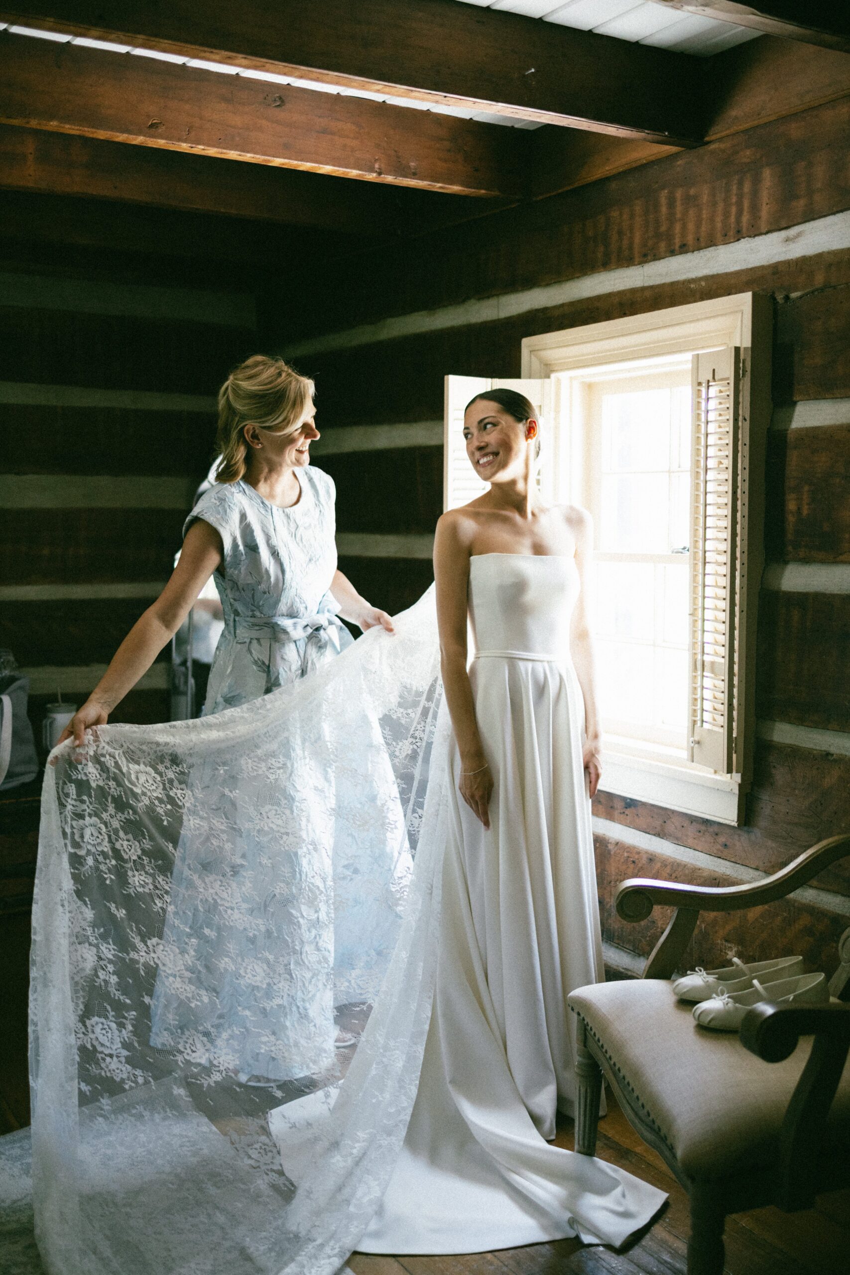 Bride smiling as a friend helps arrange her lace veil beside a sunlit window inside a rustic space at one of the beautiful wedding chapels in Nashville TN.