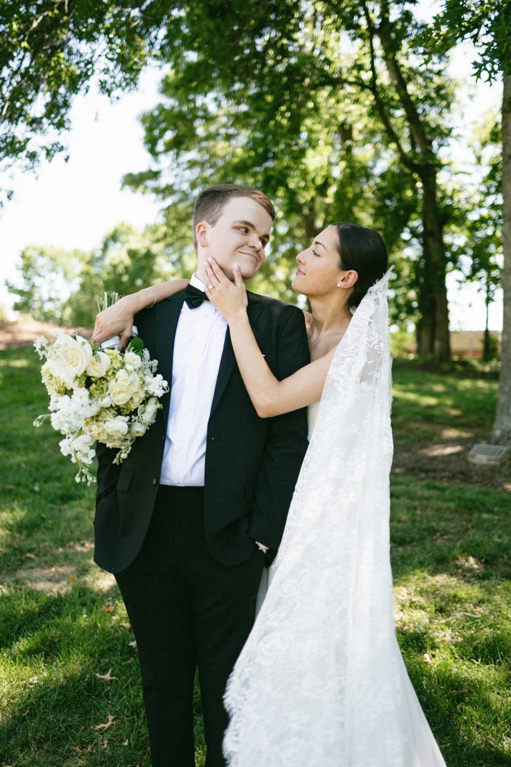 Bride gently touching the groom’s face while holding a bouquet during romantic portraits beneath tall trees at one of the most beautiful small wedding venues in Nashville TN.